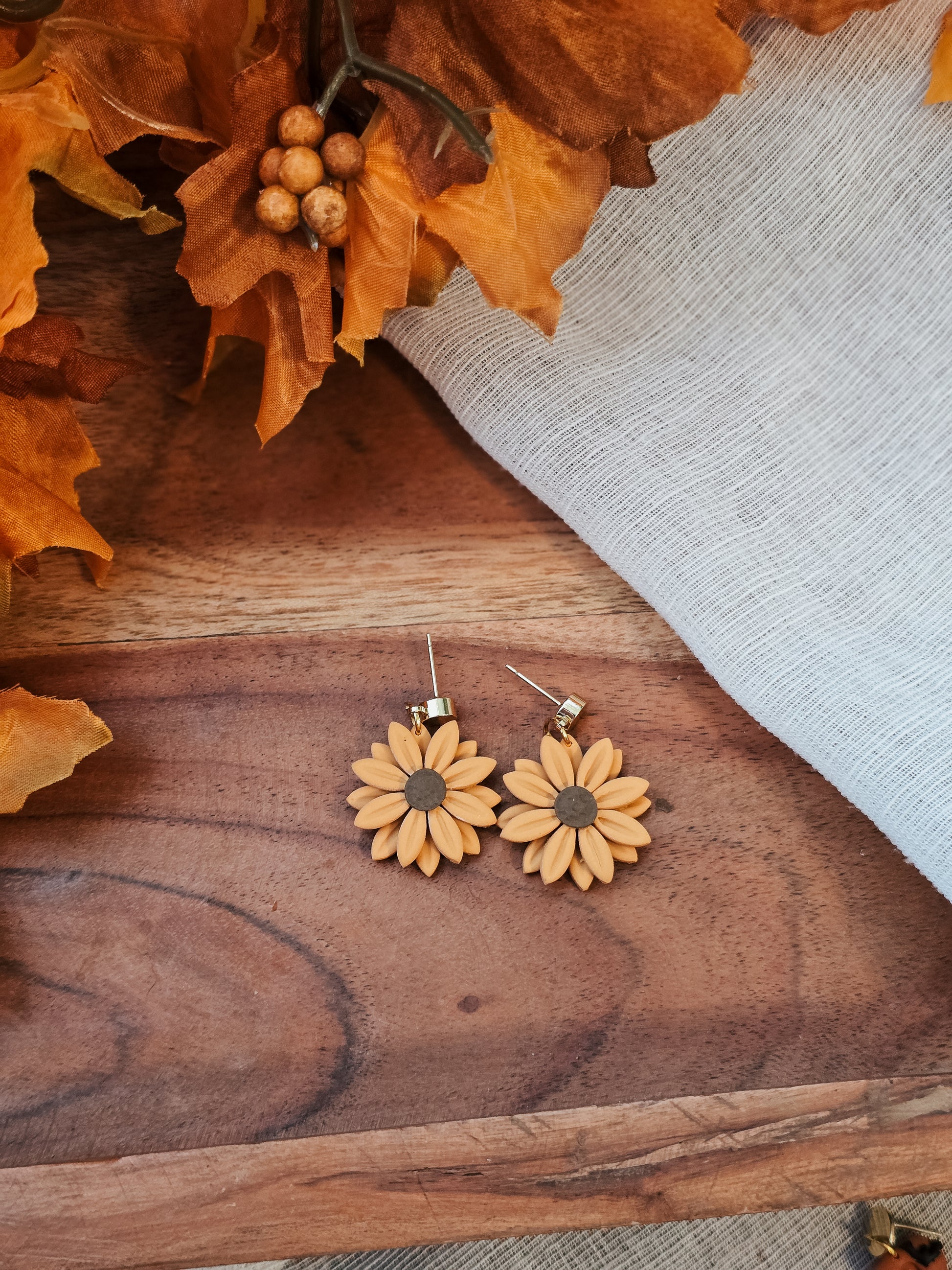 Sunflower-shaped handmade polymer clay earrings on a wooden surface with autumn leaves.